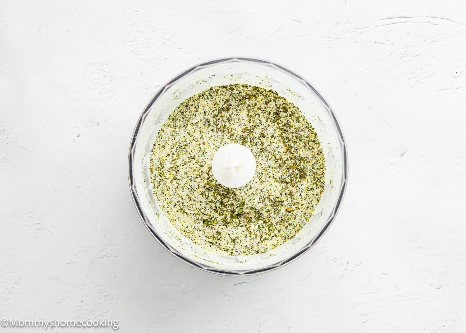 A food processor bowl filled with a finely blended green herb mixture for Homemade Ranch Seasoning Mix (Allergy-Friendly & Dairy-Free Option!) sits on a white textured surface.