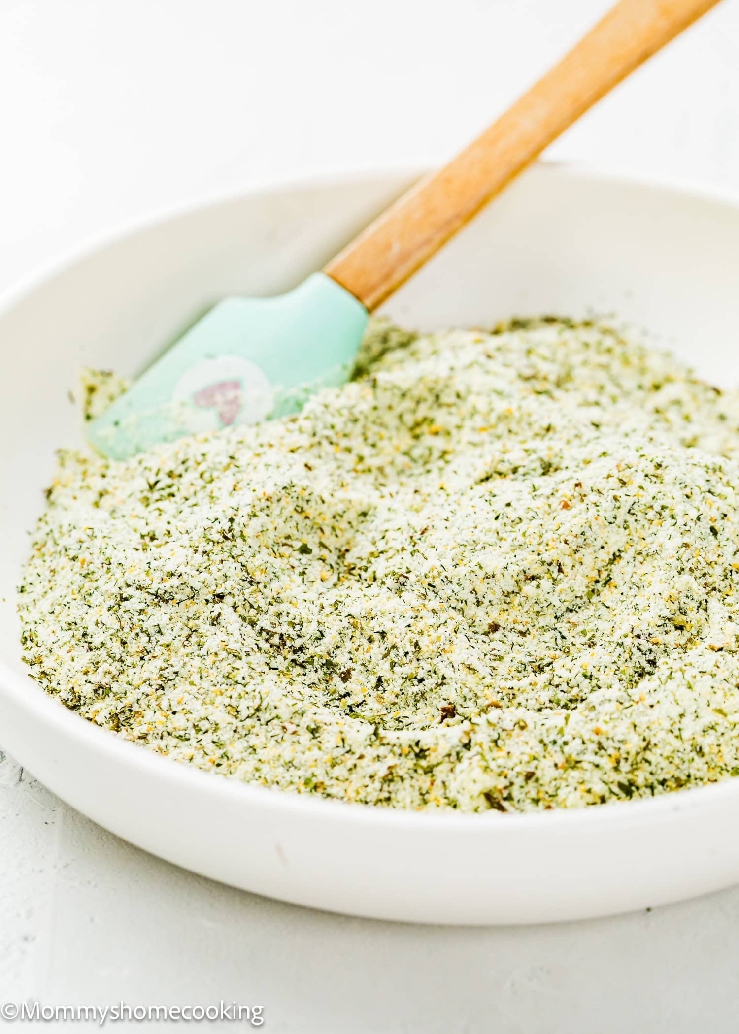 A white bowl filled with Homemade Ranch Seasoning Mix (Allergy-Friendly & Dairy-Free Option!), featuring a green and white blend, with a light blue spatula resting inside.