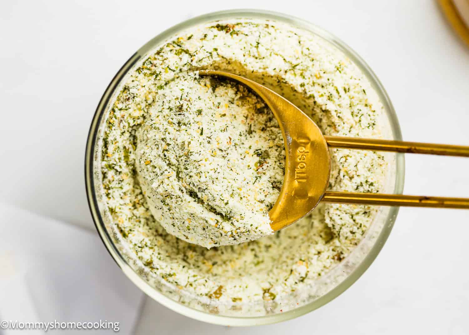A close-up of a glass bowl filled with Homemade Ranch Seasoning Mix (Allergy-Friendly & Dairy-Free Option!), with a gold measuring spoon scooping some out. Green-speckled and dry, this blend is perfect for adding flavor to your favorite dishes.