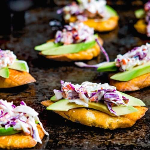 A close-up of several tostones (crispy fried green plantains) topped with sliced avocado and creamy coleslaw on a dark baking sheet.