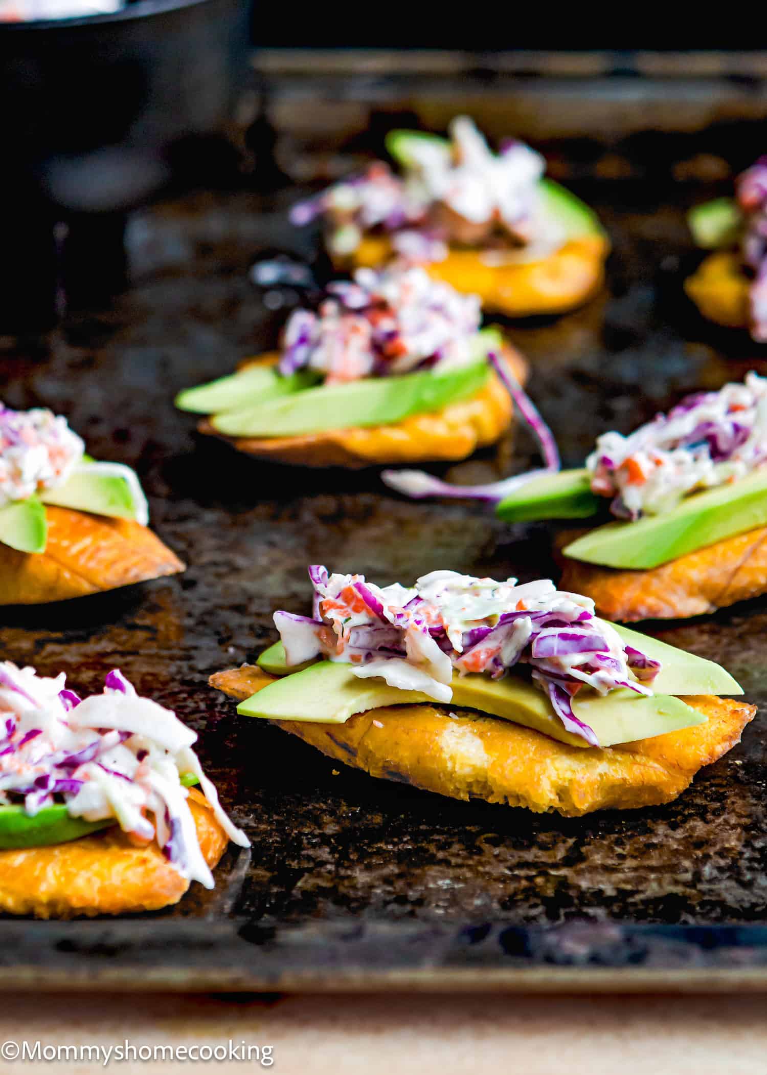 A close-up of several tostones (crispy fried green plantains) topped with sliced avocado and creamy coleslaw on a dark baking sheet.