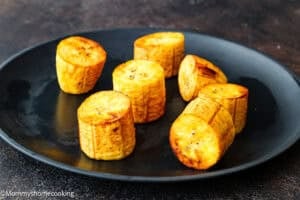 Six pieces of roasted plantain arranged on a black plate, with a dark background, showcasing the texture and appeal of How to Make Tostones (Crispy Fried Green Plantains – Easy & Authentic!).