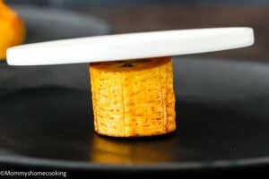 A round slice of cooked plantain stands upright on a black plate, partially covered by a white circular object, showing how to make tostones (crispy fried green plantains – easy & authentic!).