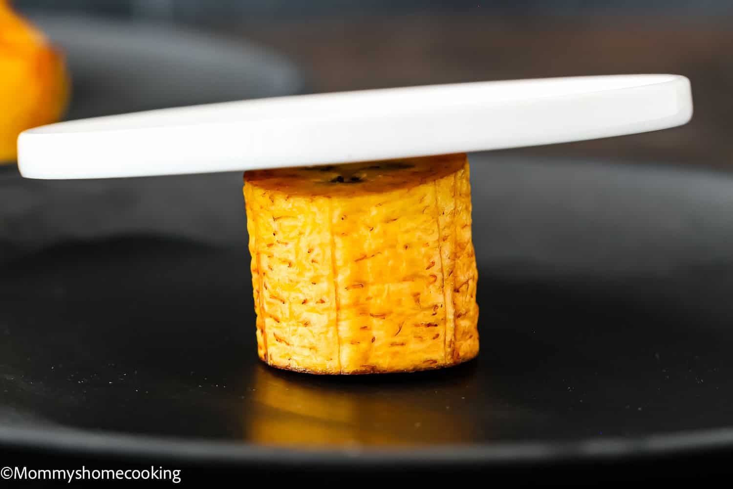 A round slice of cooked plantain stands upright on a black plate, partially covered by a white circular object, showing how to make tostones (crispy fried green plantains – easy & authentic!).