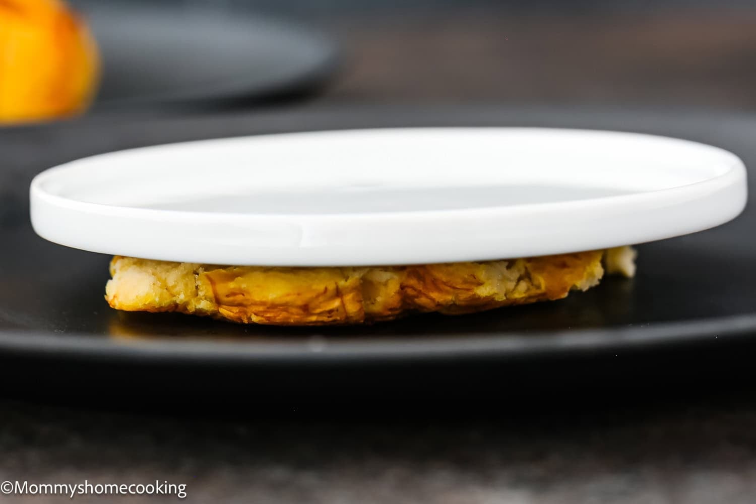 A white plate is pressing down on a biscuit or pastry on a black surface, similar to the first step in How to Make Tostones (Crispy Fried Green Plantains – Easy & Authentic!).