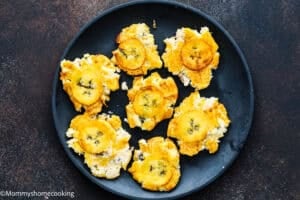 A black plate holds several pieces of smashed, fried plantains on a dark surface—showcasing how to make tostones (crispy fried green plantains) that are easy and authentic.