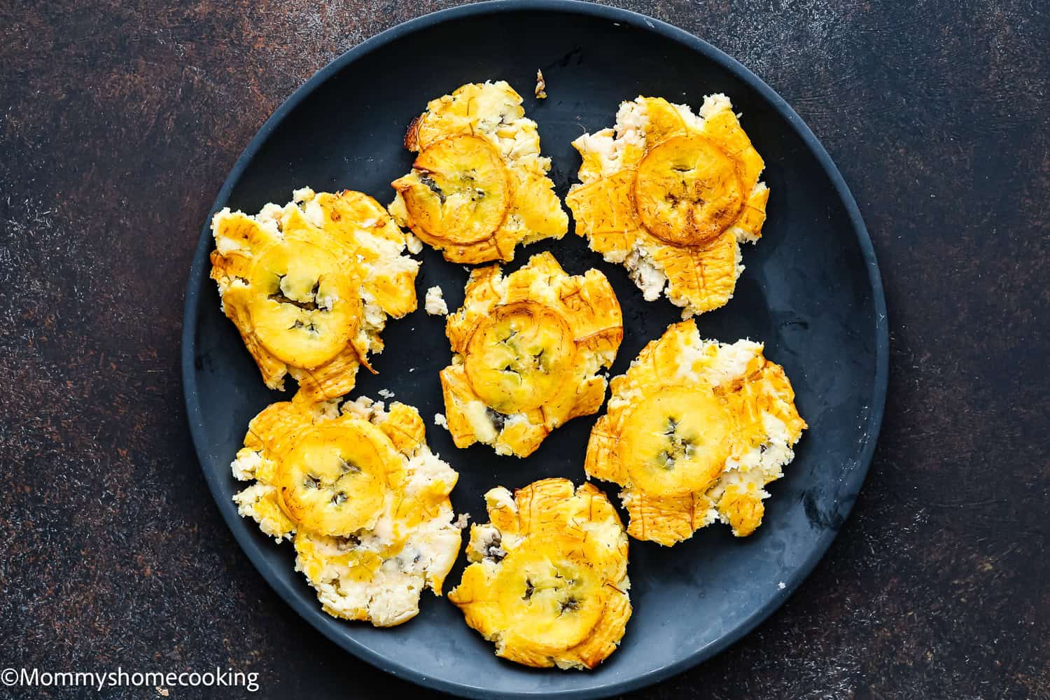 A black plate holds several pieces of smashed, fried plantains on a dark surface—showcasing how to make tostones (crispy fried green plantains) that are easy and authentic.