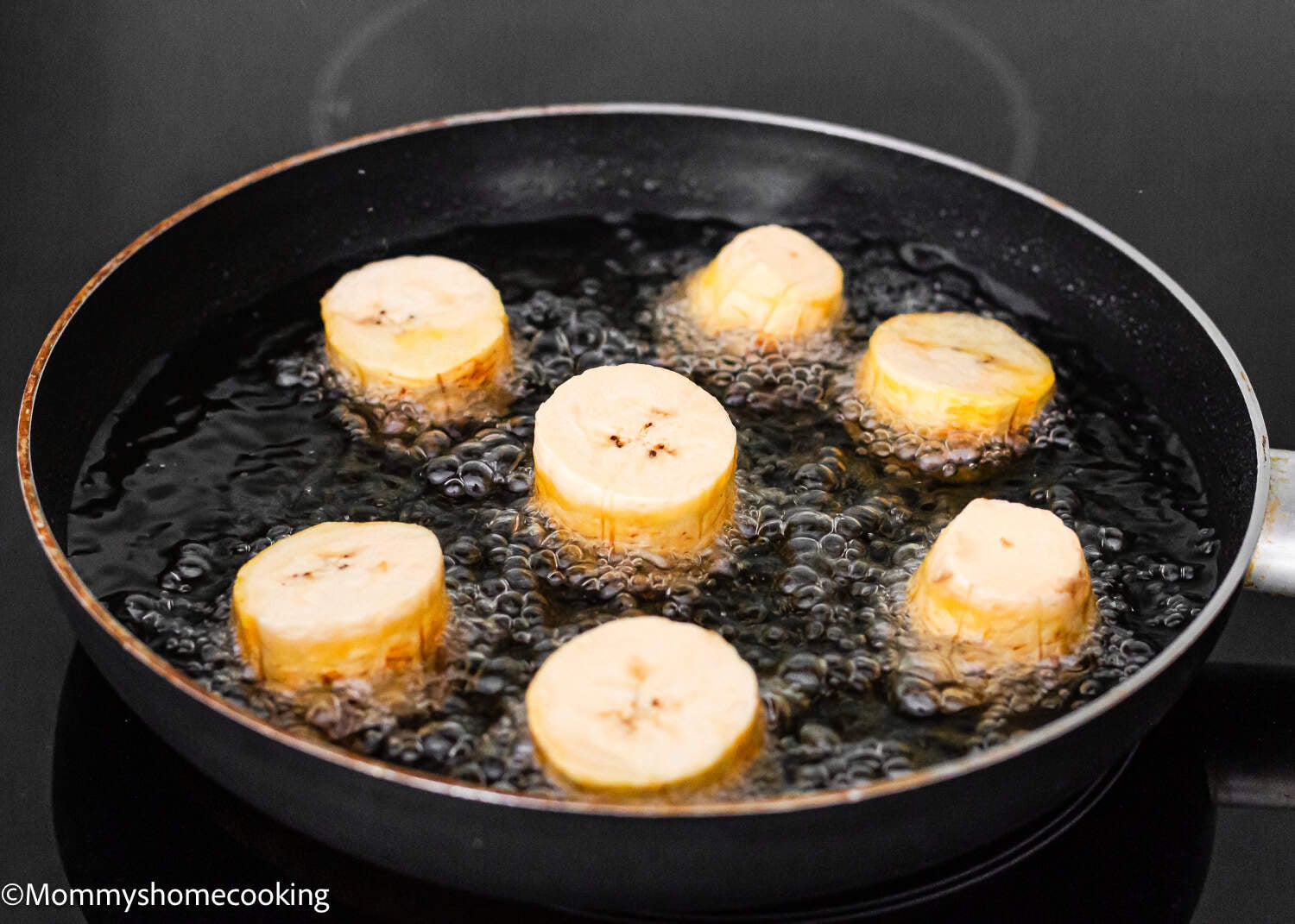 How to Make Tostones (Crispy Fried Green Plantains – Easy & Authentic!): Sliced plantains frying in oil in a black pan on a stovetop.
