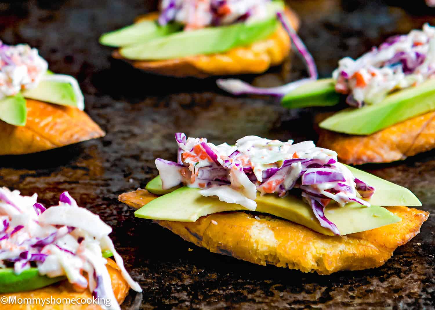 Several tostones topped with sliced avocado and a colorful coleslaw mixture are arranged on a dark surface, showing how to make tostones (crispy fried green plantains—easy & authentic!).