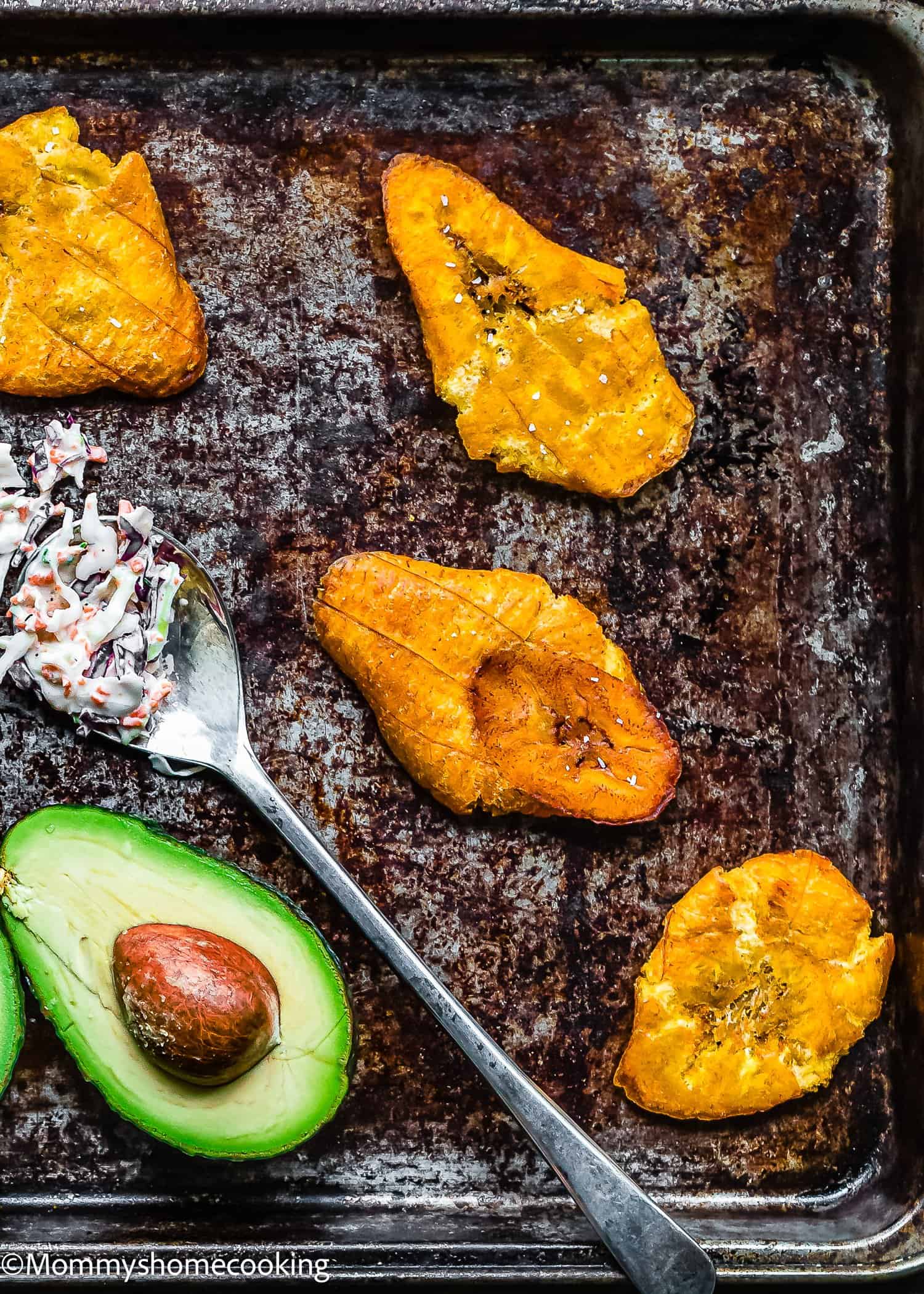 Four crispy fried plantain slices (tostones) on a baking sheet, with a halved avocado, a spoonful of creamy salad, and a pit visible in the lower left corner. Learn How to Make Tostones (Crispy Fried Green Plantains – Easy & Authentic!).