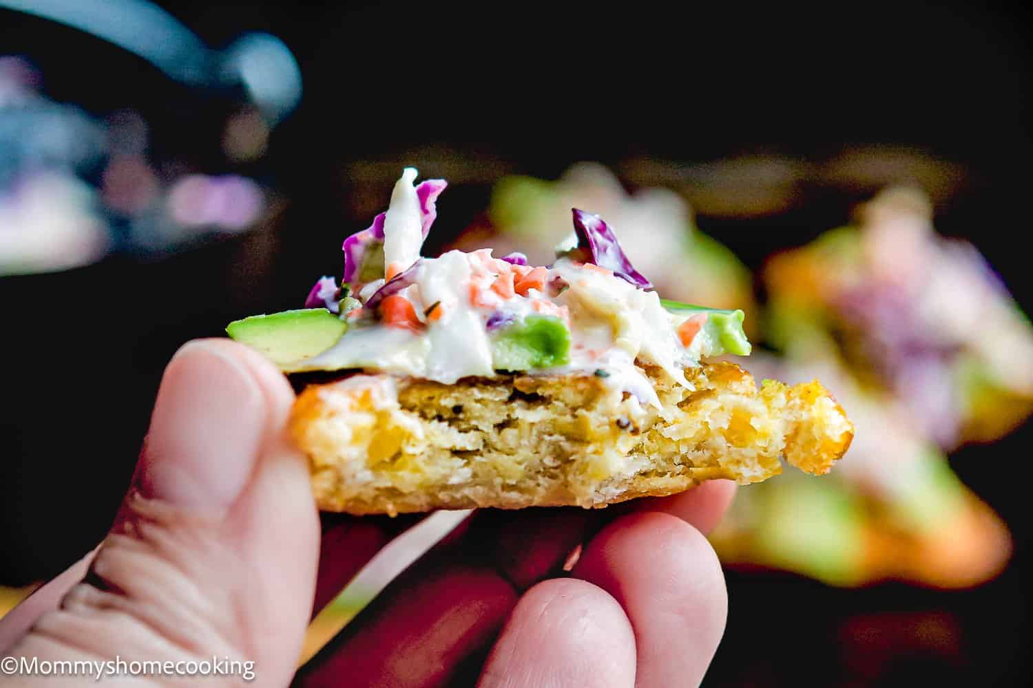 A hand holds a partially eaten fried patty topped with coleslaw, avocado, and vegetables, similar to How to Make Tostones (Crispy Fried Green Plantains – Easy & Authentic!).