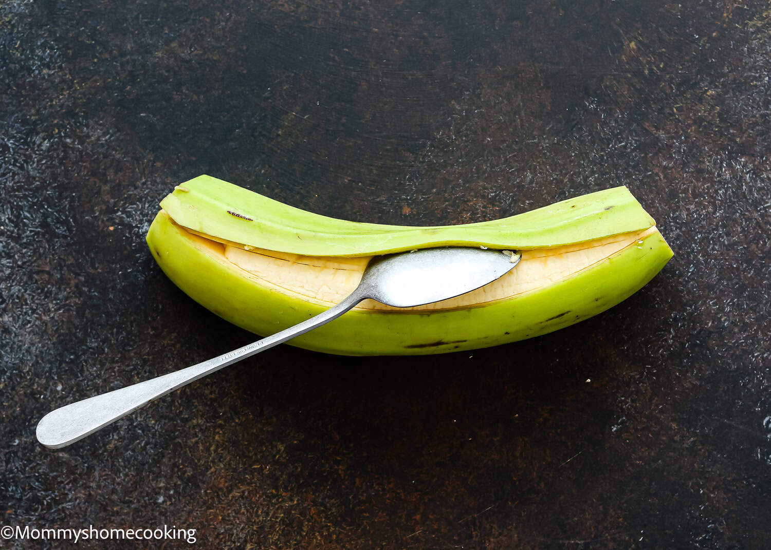 A green plantain with a lengthwise slit is shown on a dark surface, a metal spoon inserted into the opening—an essential step in How to Make Tostones (Crispy Fried Green Plantains – Easy & Authentic!).