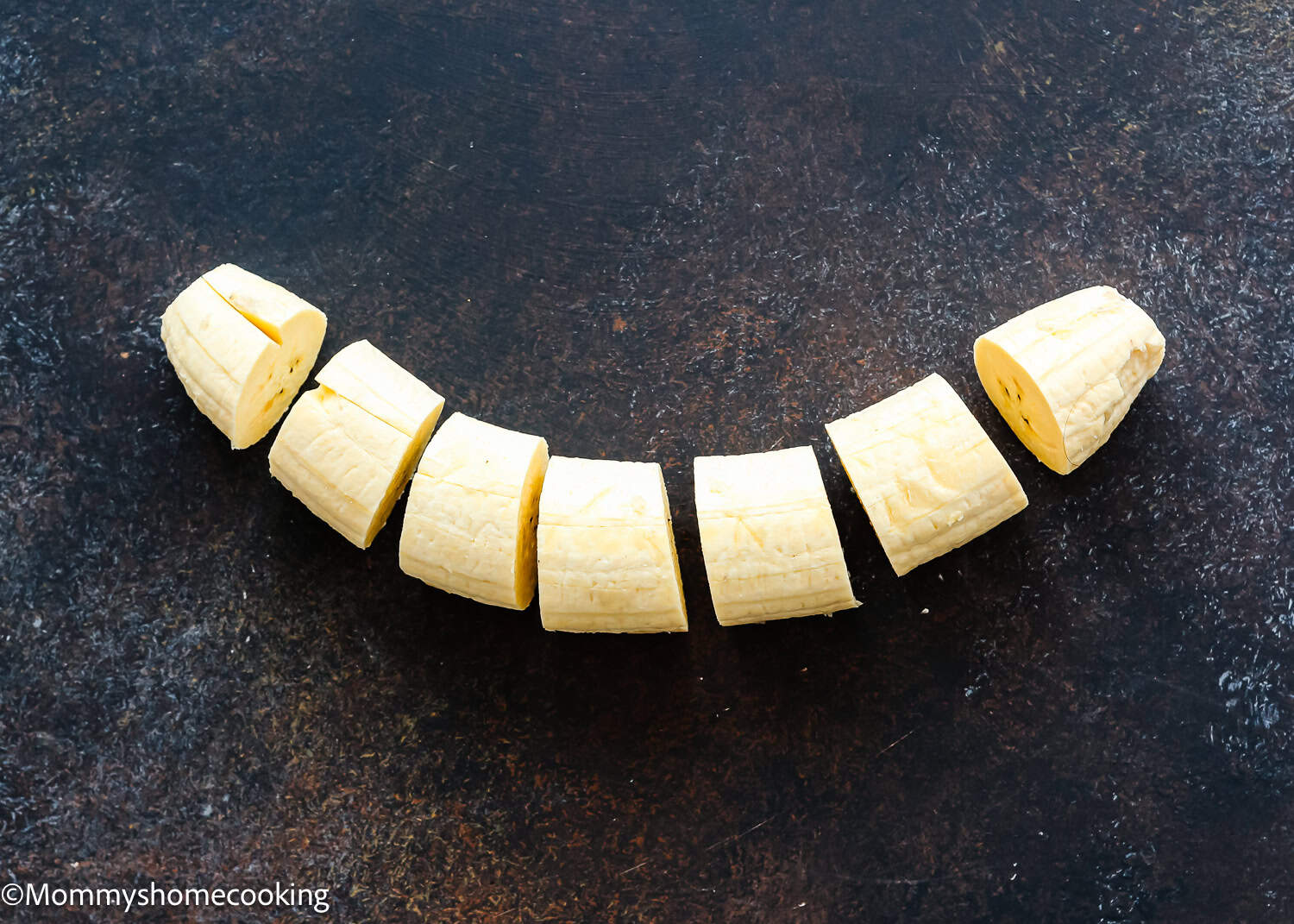 Sliced pieces of peeled plantain arranged in a row on a dark, textured surface—perfect for learning How to Make Tostones (Crispy Fried Green Plantains – Easy & Authentic!).