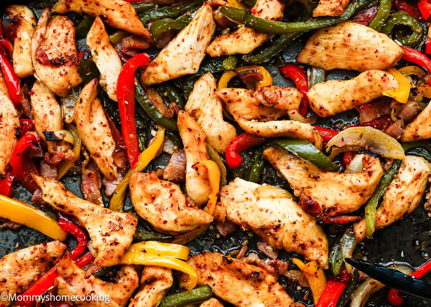 Sheet Pan Barbecue Bacon Chicken Fajitas featuring cooked chicken strips with red, yellow, and green bell peppers and onions, all seasoned with spices on a dark surface.