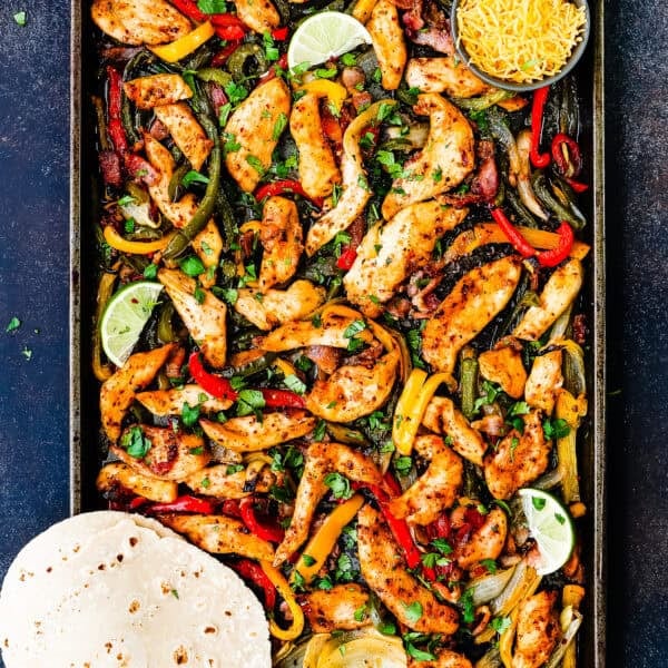 A sheet pan filled with Sheet Pan Barbecue Bacon Chicken Fajitas, bell peppers, onions, lime wedges, shredded cheese, and a stack of flour tortillas on the side.