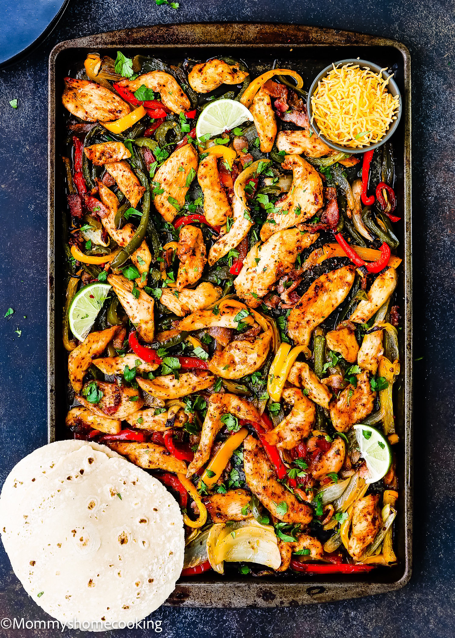 A sheet pan filled with Sheet Pan Barbecue Bacon Chicken Fajitas, bell peppers, onions, lime wedges, shredded cheese, and a stack of flour tortillas on the side.