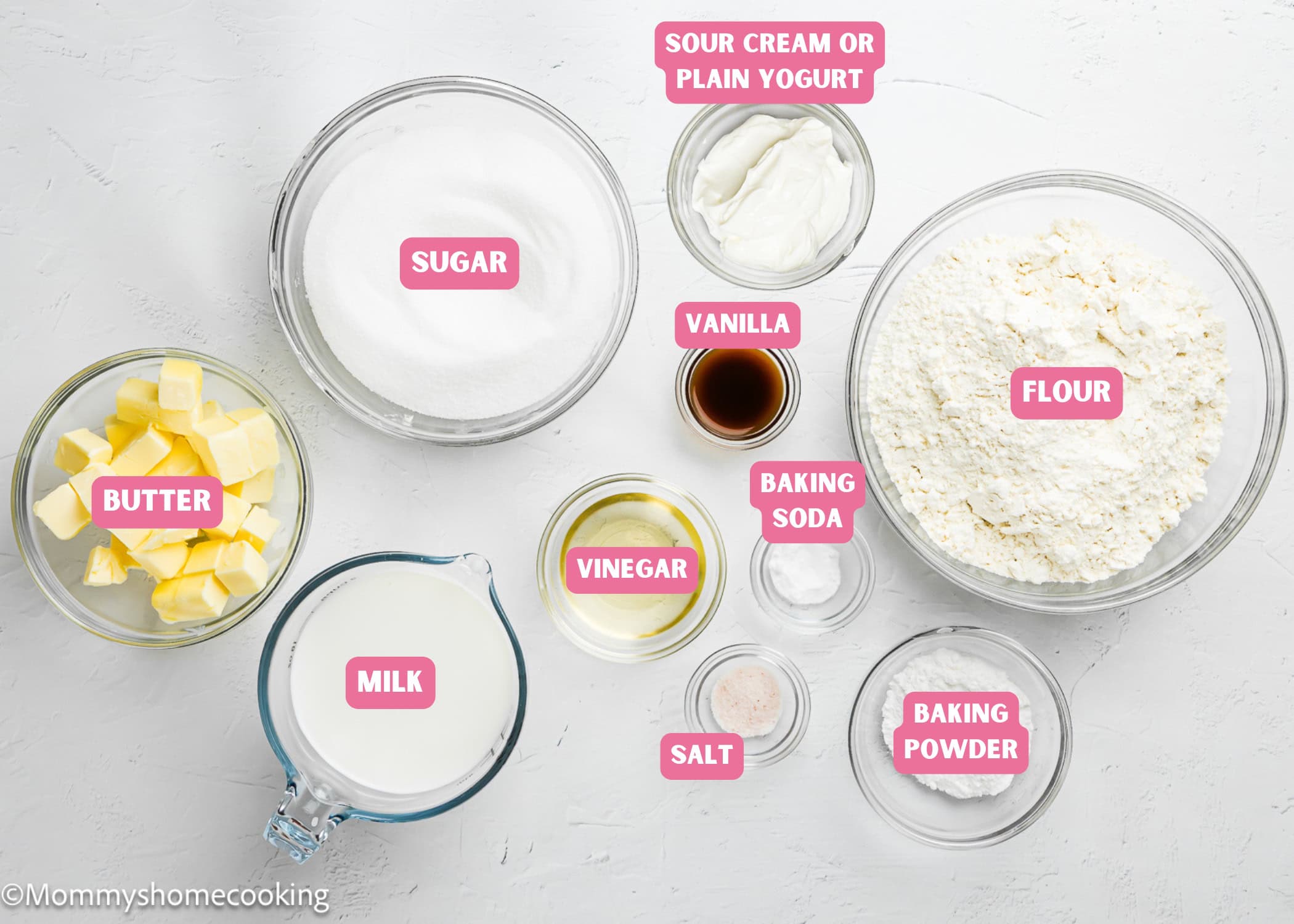 Bowls containing butter cubes, sugar, flour, milk, sour cream, vanilla extract, oil, baking powder, salt, and cornstarch are arranged on a white surface—perfect for making a soft, fluffy Egg-Free Vanilla Cake.