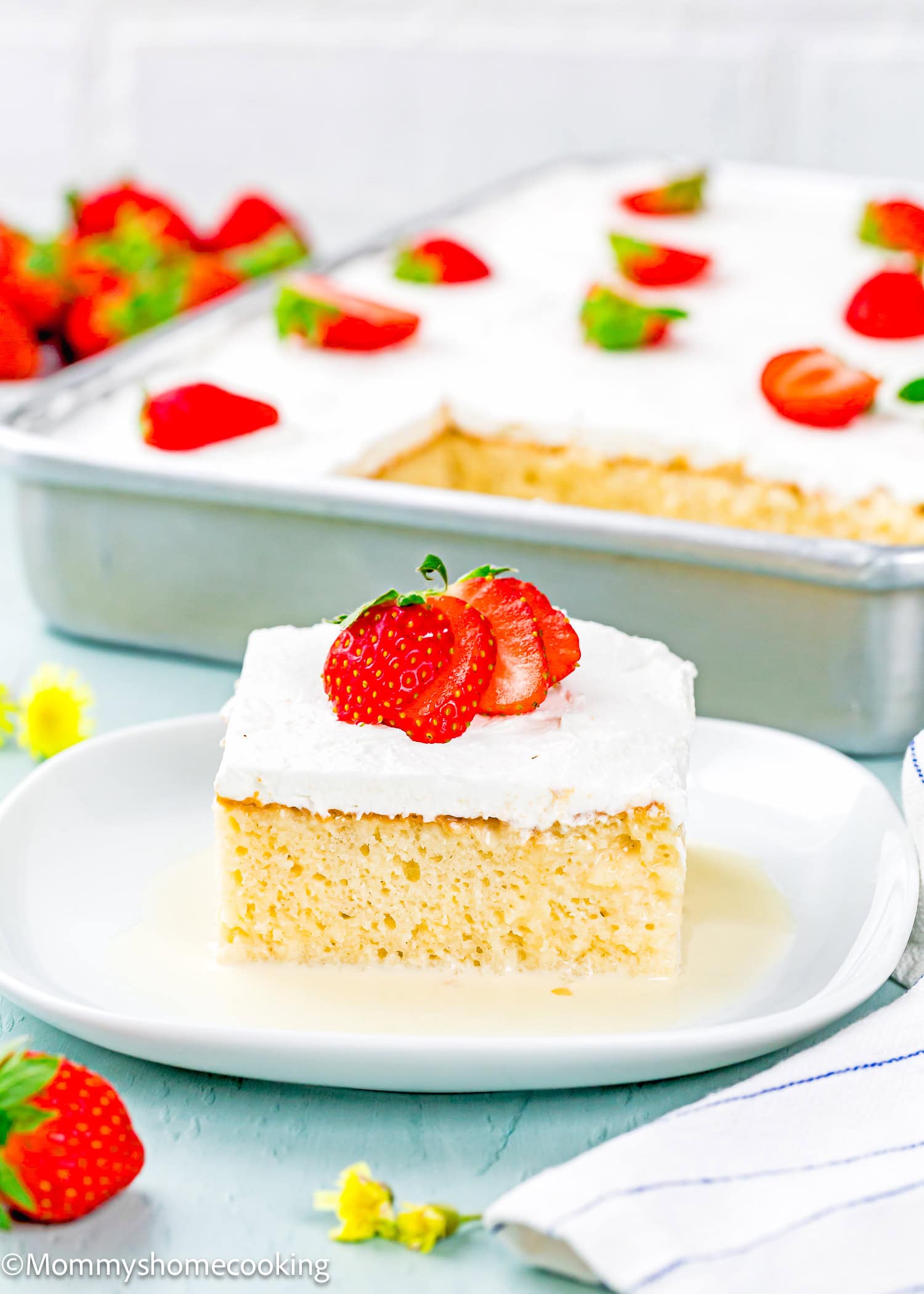 A slice of The Best Eggless Tres Leches Cake topped with whipped cream and sliced strawberries sits on a white plate, with the remaining cake in a pan in the background.