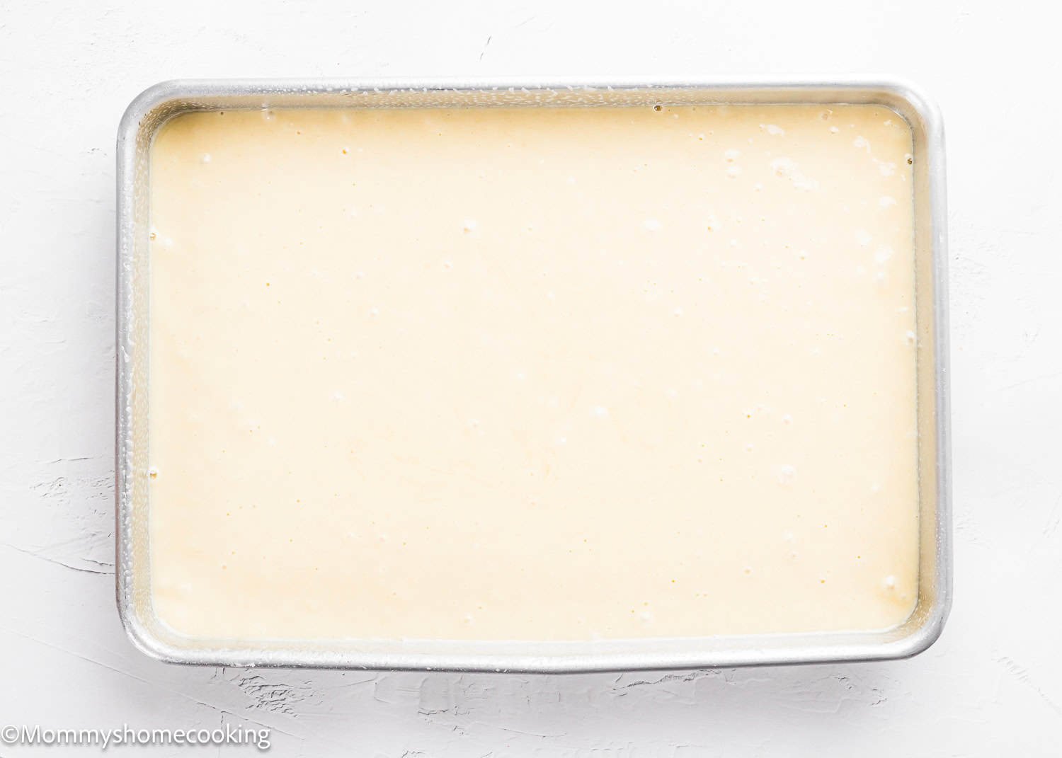 A rectangular metal baking pan filled with smooth, light-colored batter for The Best Eggless Tres Leches Cake sits on a white surface.