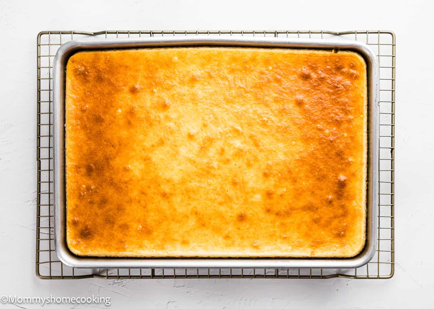 A rectangular cheesecake in a metal baking pan rests on a cooling rack against a white background, similar to The Best Eggless Tres Leches Cake.