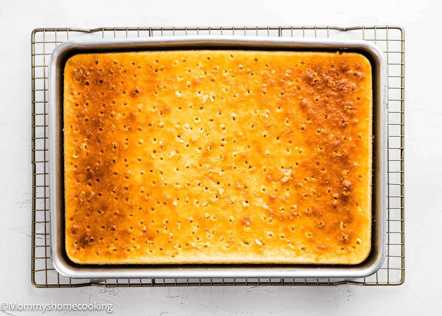 A rectangular sheet cake, The Best Eggless Tres Leches Cake, with small holes poked across the surface sits in a metal baking pan on a cooling rack.