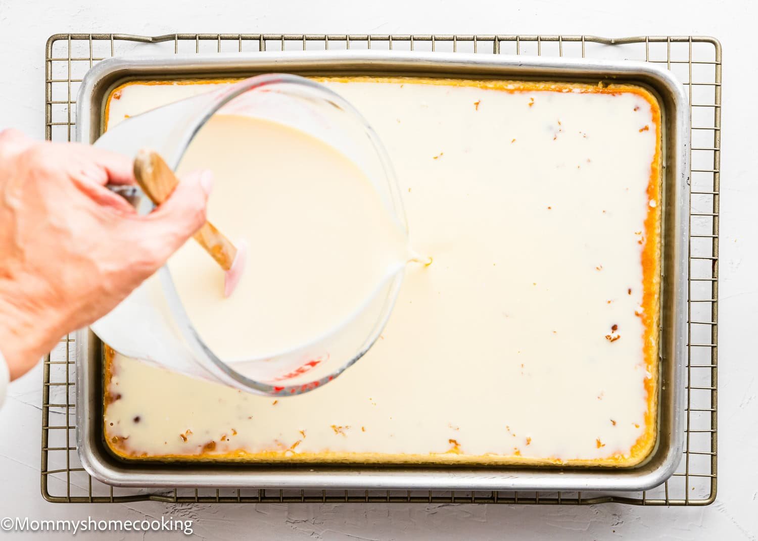 A hand pours a creamy liquid from a measuring cup onto The Best Eggless Tres Leches Cake in a rectangular pan set on a cooling rack.