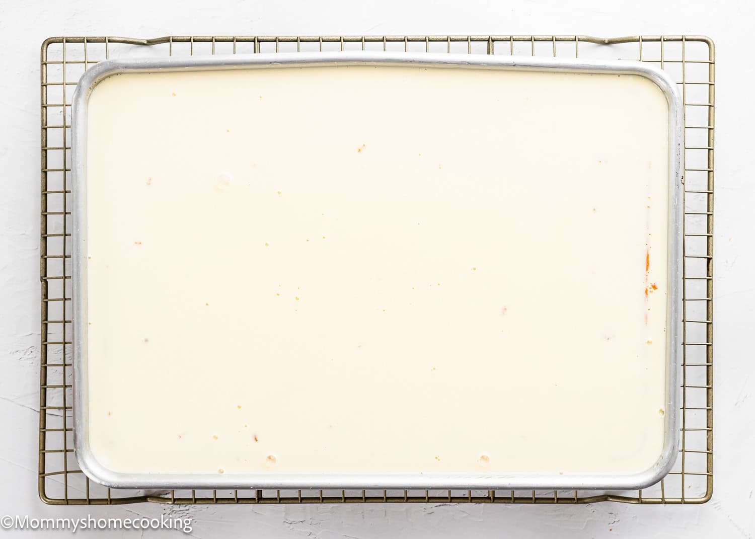 A rectangular metal baking pan filled with a smooth, light cream-colored mixture for The Best Eggless Tres Leches Cake sits on a wire rack over a white surface.