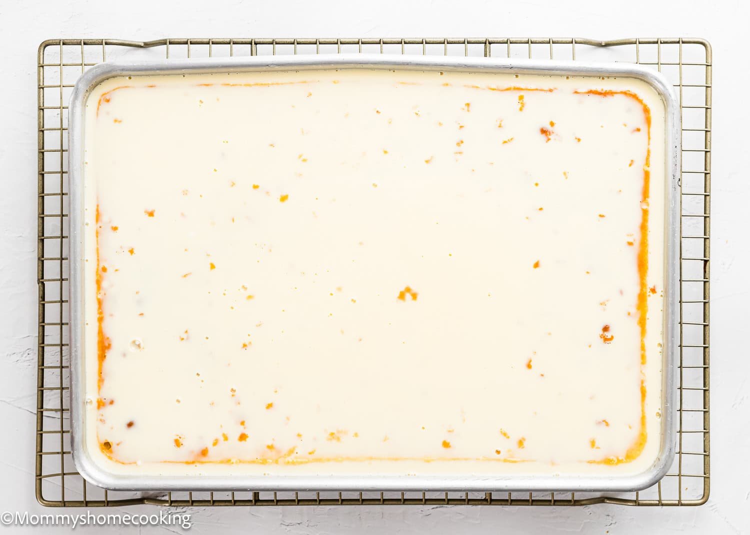 A rectangular baking tray filled with a creamy white dessert, possibly The Best Eggless Tres Leches Cake with milk poured over it, rests on a wire cooling rack.