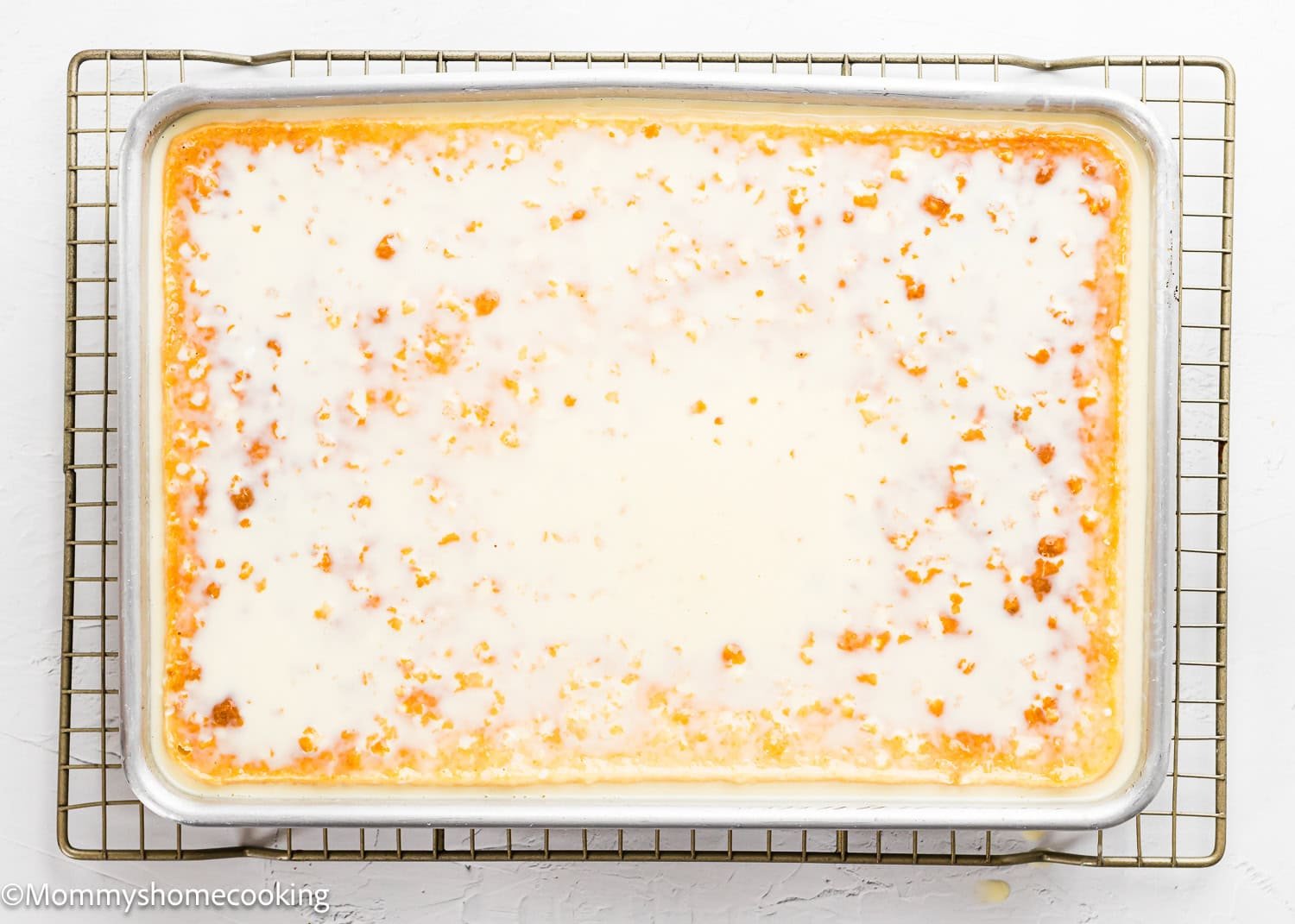 A rectangular sheet cake with white glaze, inspired by The Best Eggless Tres Leches Cake, sits on a cooling rack over a baking tray.
