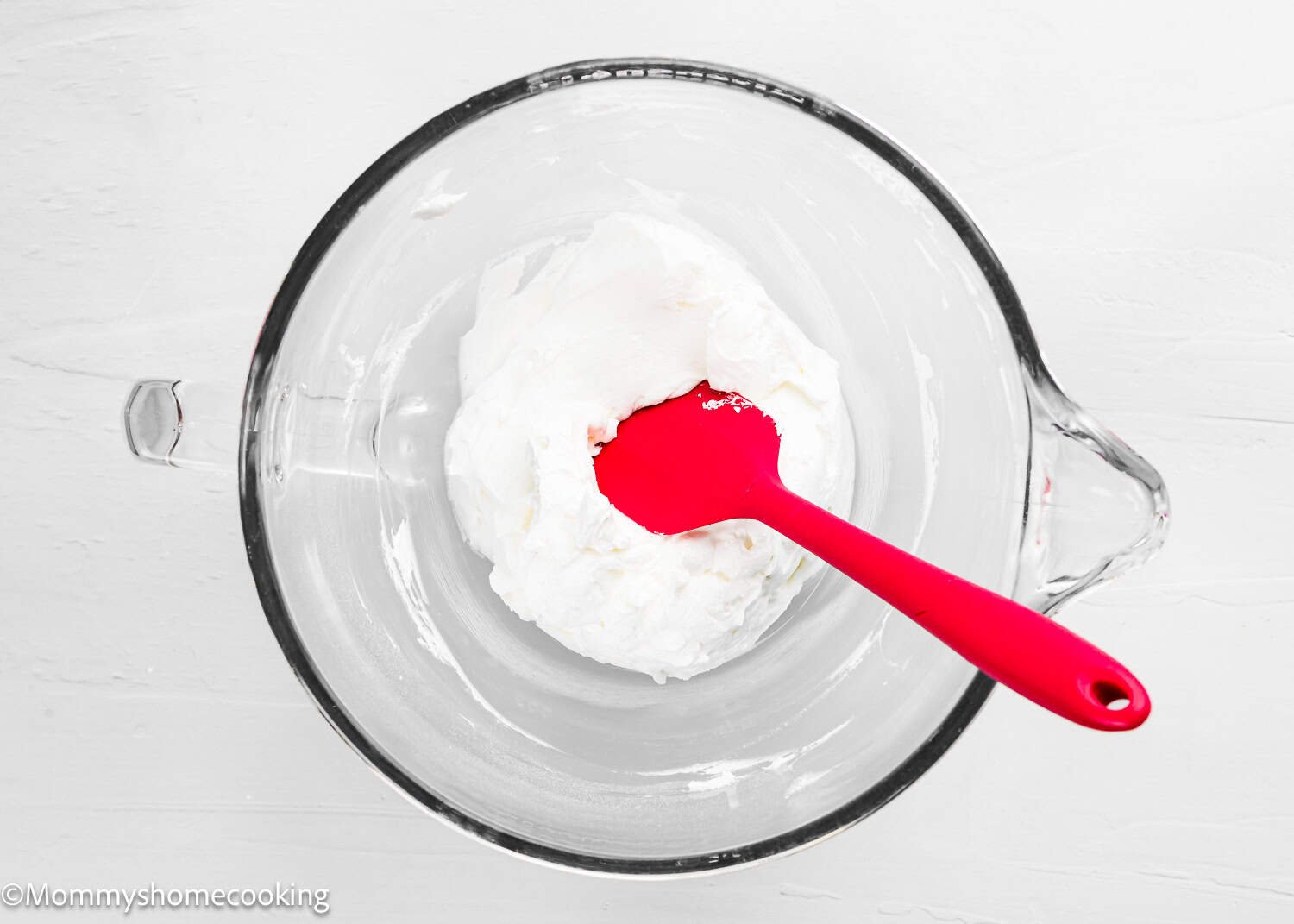 A glass bowl containing a white creamy mixture for The Best Eggless Tres Leches Cake, with a red spatula resting inside.