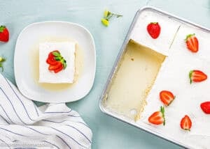 A square slice of The Best Eggless Tres Leches Cake topped with strawberries sits on a plate next to a baking pan of cake, with a striped napkin nearby.