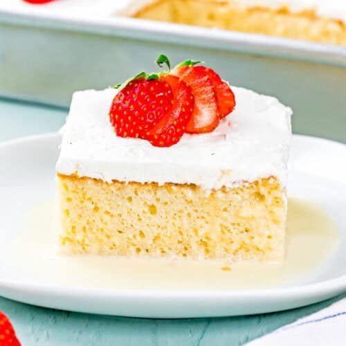 A slice of The Best Eggless Tres Leches Cake topped with whipped cream and sliced strawberries sits on a white plate, with the remaining cake in a pan in the background.