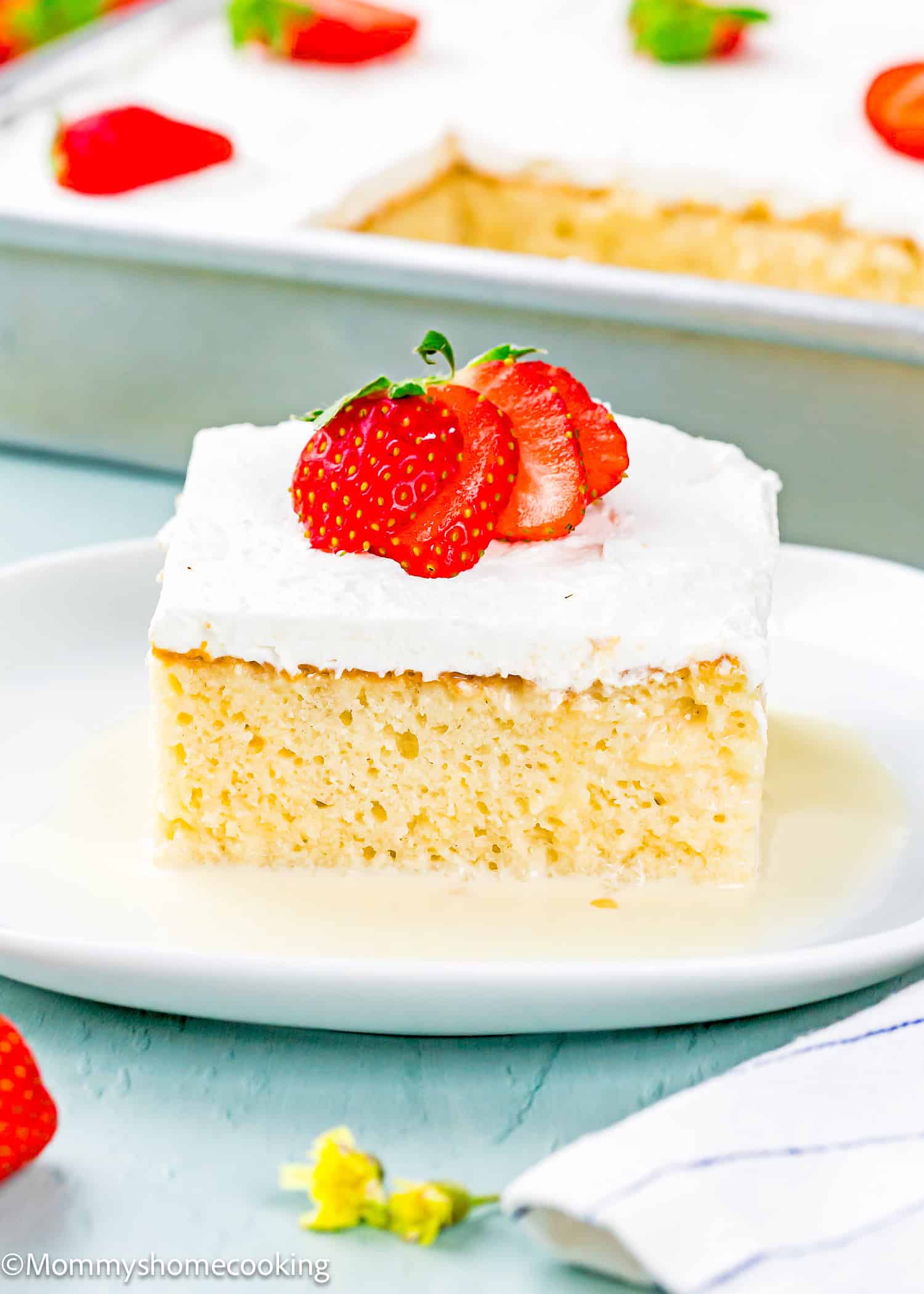 A slice of The Best Eggless Tres Leches Cake topped with whipped cream and sliced strawberries sits on a white plate, with the remaining cake in a pan in the background.