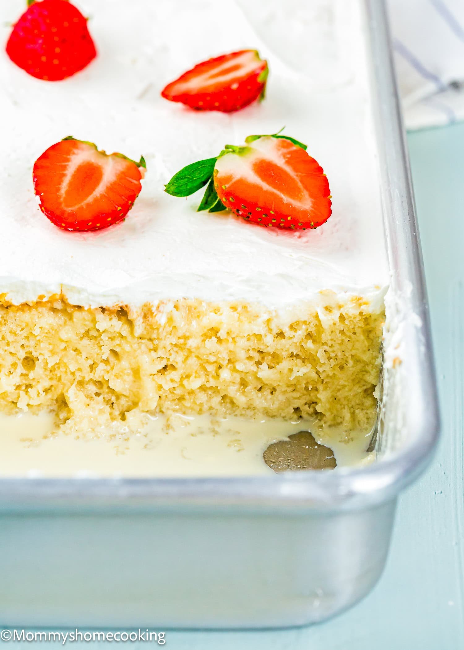 A rectangular cake with white frosting is topped with halved strawberries. A portion is cut out, revealing the moist interior and liquid pooled at the bottom—truly The Best Eggless Tres Leches Cake.