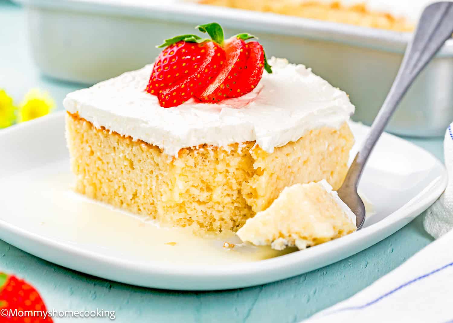 A slice of The Best Eggless Tres Leches Cake with white frosting, topped with sliced strawberries, sits on a white plate with a forkful taken out.