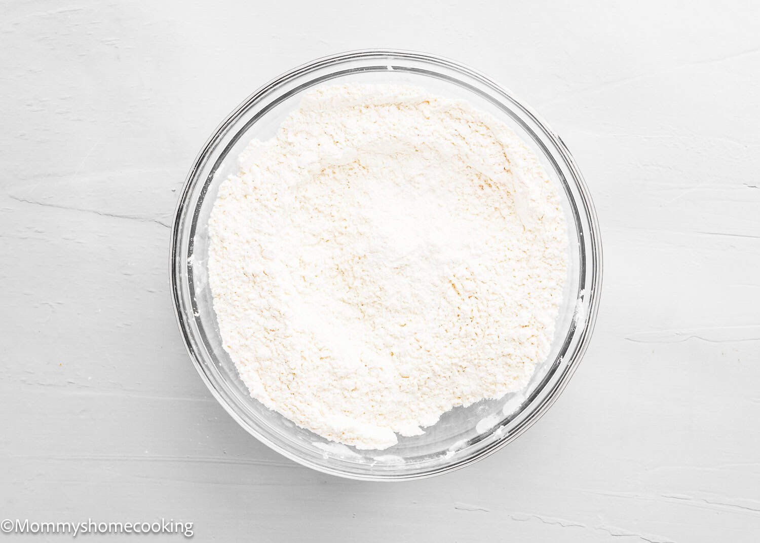 A clear glass bowl filled with white flour sits on a white surface, perfect for preparing The Best Eggless Tres Leches Cake.