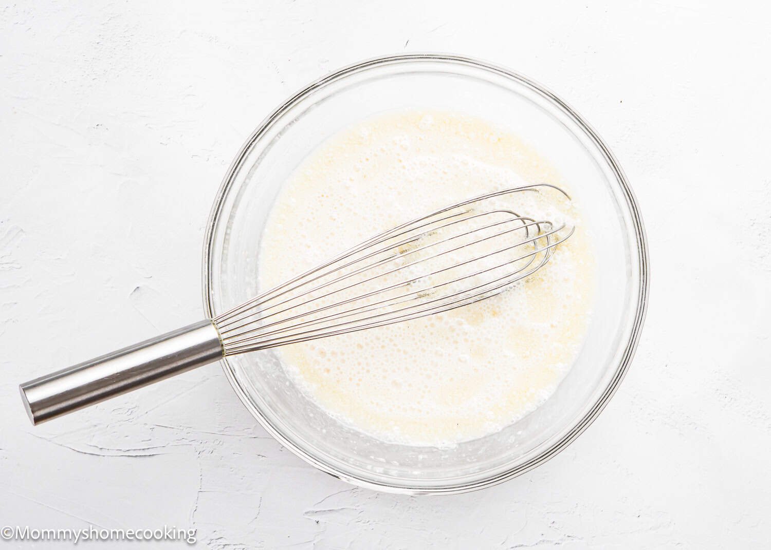 A metal whisk rests in a glass bowl containing a light, frothy mixture on a white surface, perfect for making The Best Eggless Tres Leches Cake.