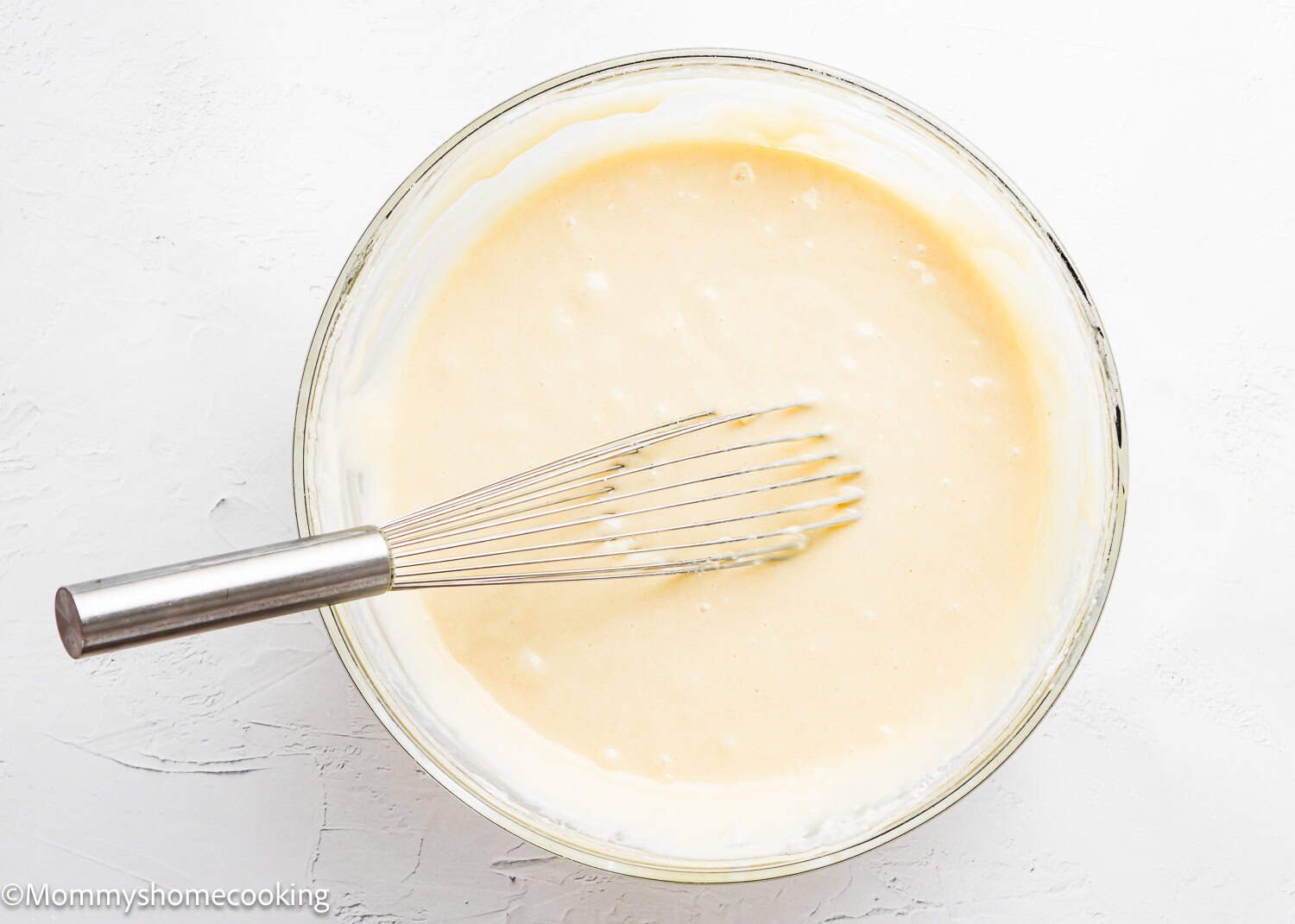 A metal whisk rests in a glass bowl filled with smooth, pale yellow batter for The Best Eggless Tres Leches Cake on a white surface.