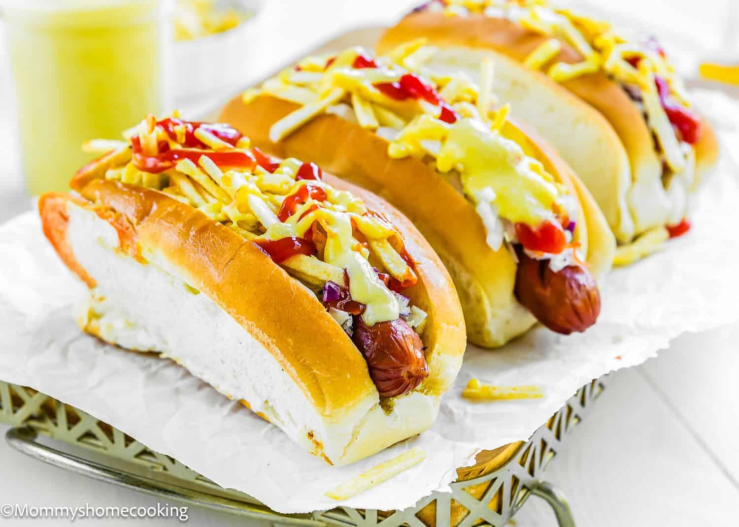 Three hot dogs in buns topped with shredded cheese, ketchup, mustard, and a drizzle of The Best Sweet Corn Sauce (Venezuelan-Style & Irresistibly Creamy), arranged on parchment paper.