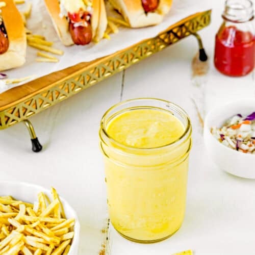 A jar of The Best Sweet Corn Sauce (Venezuelan-Style & Irresistibly Creamy) sits on a white table next to a bowl of thin fries, a bowl of coleslaw, and hot dogs topped with sauce on a tray.