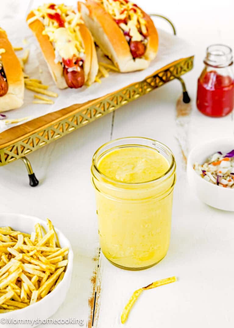 A jar of The Best Sweet Corn Sauce (Venezuelan-Style & Irresistibly Creamy) sits on a white table next to a bowl of thin fries, a bowl of coleslaw, and hot dogs topped with sauce on a tray.