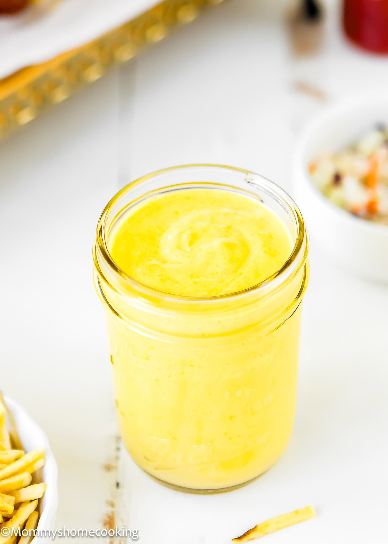 A glass jar filled with The Best Sweet Corn Sauce (Venezuelan-Style & Irresistibly Creamy) sits on a white table next to a bowl of fries and a small bowl of coleslaw.