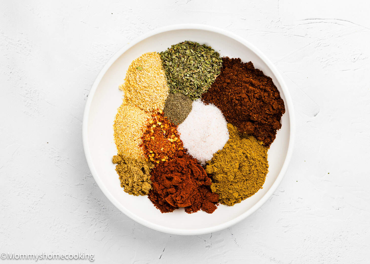 A white bowl contains separate piles of various dried spices and seasonings, including The Best Taco Seasoning Recipe (Allergy-Safe & Family-Friendly), arranged in a circular pattern on a white surface.