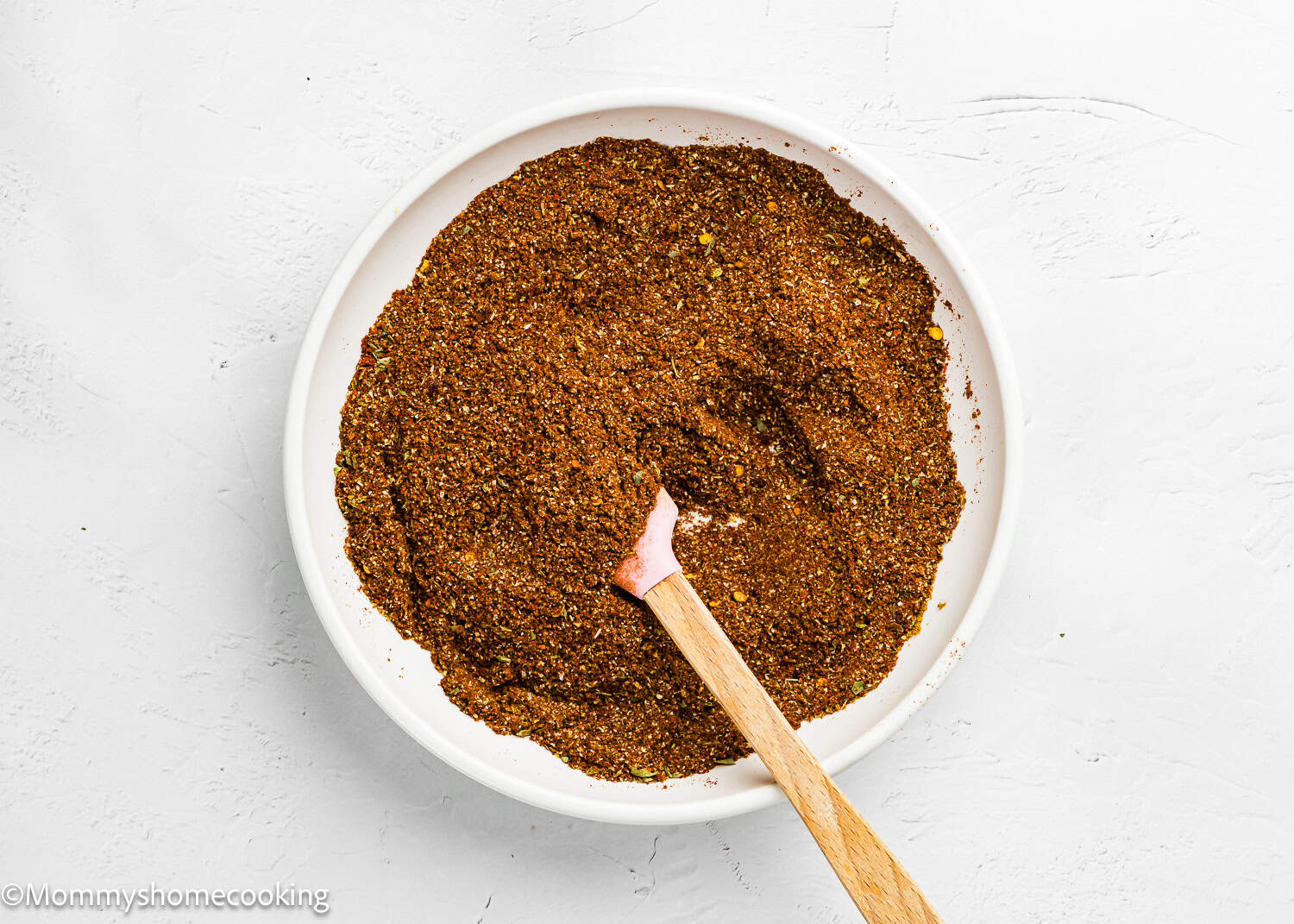 A white bowl filled with a dark brown spice mix—the Best Taco Seasoning Recipe (Allergy-Safe & Family-Friendly)—with a wooden spoon resting inside, on a light textured surface.