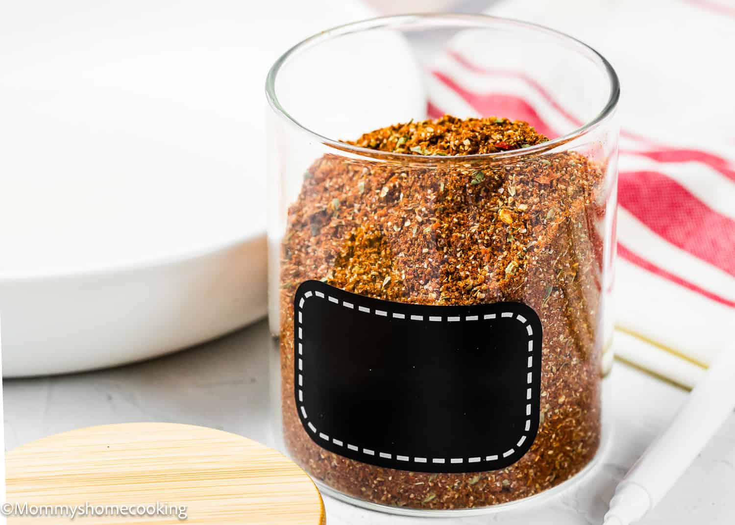 A glass jar filled with a reddish-brown spice blend sits on a white surface, featuring a blank black label. A wooden lid and a red-striped towel are nearby, showcasing The Best Taco Seasoning Recipe (Allergy-Safe & Family-Friendly).