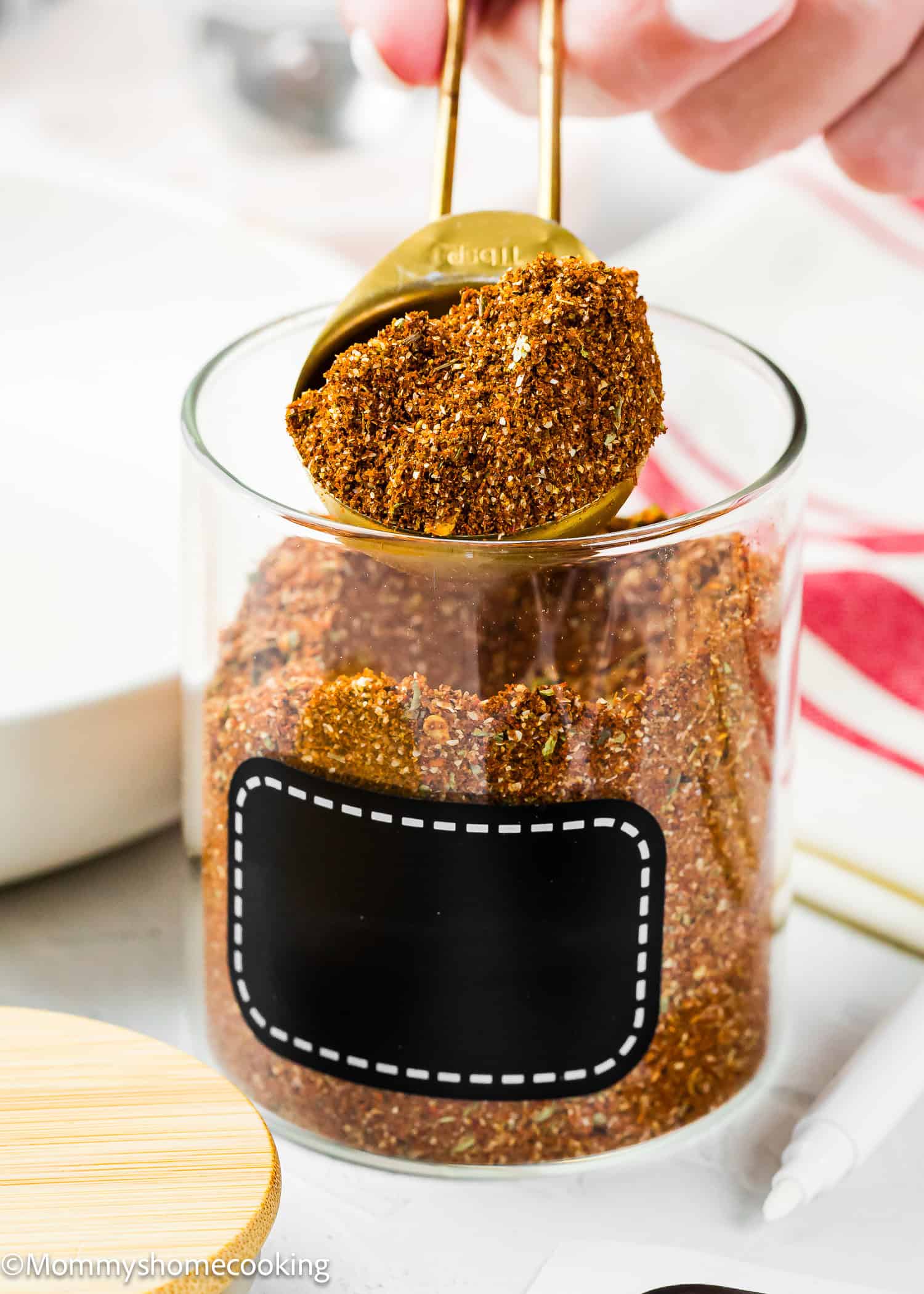 A hand scoops a spoonful of The Best Taco Seasoning Recipe (Allergy-Safe & Family-Friendly) from a glass jar with a blank black label.