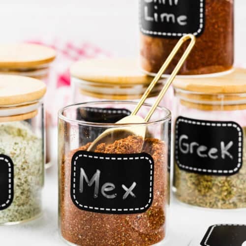 Glass jars with wooden lids are labeled "Mex," "Greek," and "Chili Lime," each filled with different seasoning blends. A gold spoon rests in the "Mex" jar, featuring The Best Taco Seasoning Recipe (Allergy-Safe & Family-Friendly).