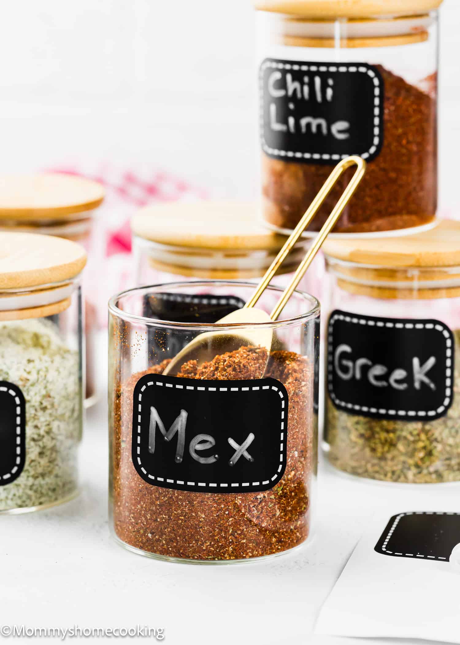 Glass jars with wooden lids are labeled "Mex," "Greek," and "Chili Lime," each filled with different seasoning blends. A gold spoon rests in the "Mex" jar, featuring The Best Taco Seasoning Recipe (Allergy-Safe & Family-Friendly).