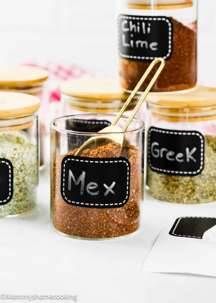 Glass jars with wooden lids are labeled "Mex," "Greek," and "Chili Lime," each filled with different seasoning blends. A gold spoon rests in the "Mex" jar, featuring The Best Taco Seasoning Recipe (Allergy-Safe & Family-Friendly).