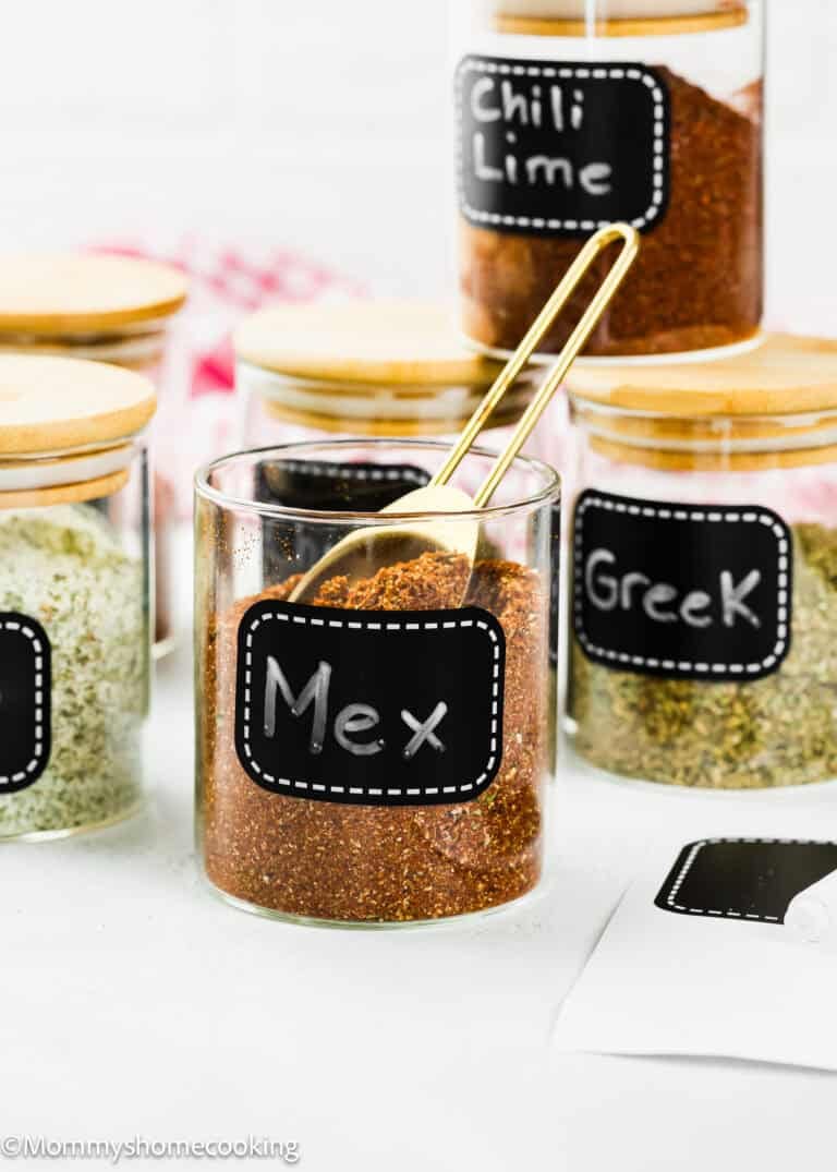 Glass jars with wooden lids are labeled "Mex," "Greek," and "Chili Lime," each filled with different seasoning blends. A gold spoon rests in the "Mex" jar, featuring The Best Taco Seasoning Recipe (Allergy-Safe & Family-Friendly).